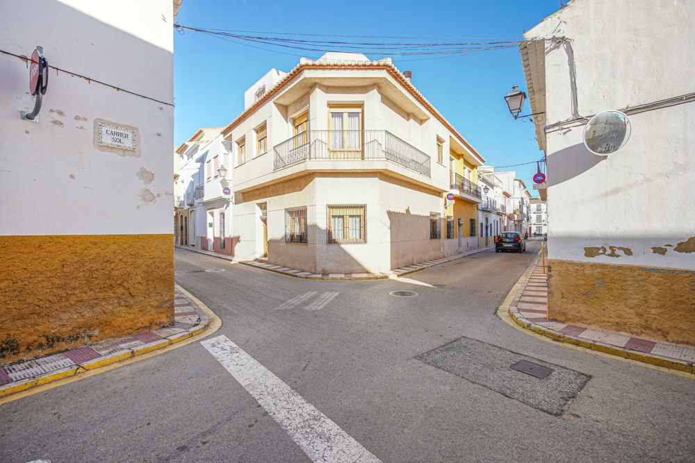 Fachada exterior de esquina de una casa de pueblo tradicional en Gata de Gorgos, con balcón de forja y ventanas protegidas en una calle tranquila y soleada.