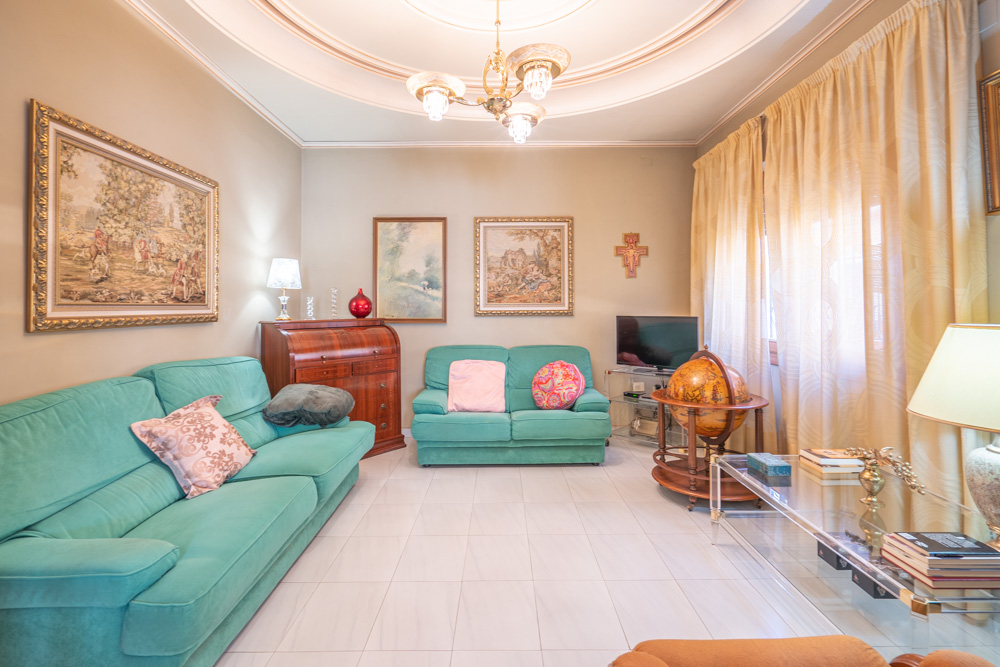 Salón luminoso con sofás turquesa, suelos de gres blanco y molduras circulares en el techo. Incluye mobiliario clásico de madera, un escritorio de persiana y cuadros decorativos.