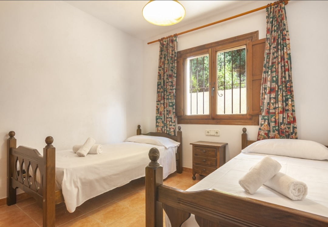 Dormitorio con dos camas individuales de madera tallada, suelos de terracota y una ventana de madera con vistas al jardín exterior.