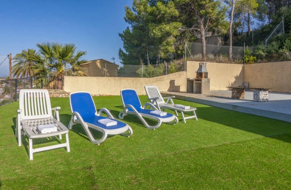 Zona de terraza con césped artificial, cuatro tumbonas, barbacoa de obra y un banco de gaviones, rodeada de pinos y vegetación mediterránea.