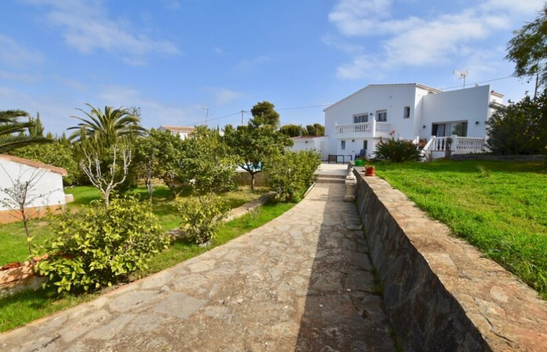 Camino de piedra natural que atraviesa el jardín mediterráneo hacia la villa blanca, rodeado de césped, palmeras y árboles frutales bajo un cielo despejado.