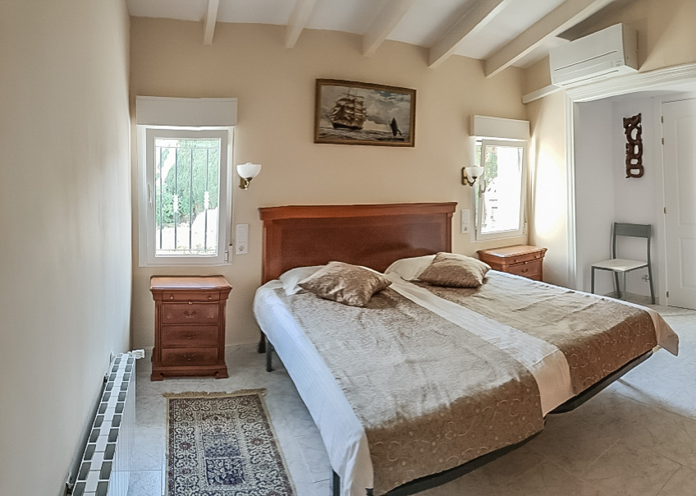 Dormitorio con techos de vigas blancas, cama doble con cabecero de madera, mesitas a juego, aire acondicionado y calefacción por radiador.