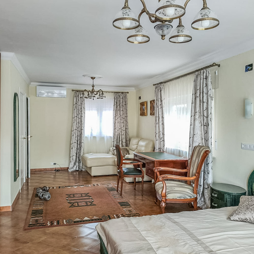 Dormitorio espacioso con zona de estar, escritorio de madera clásica, aire acondicionado y suelos de gres. Grandes ventanales con cortinas estampadas aportan abundante luz natural.