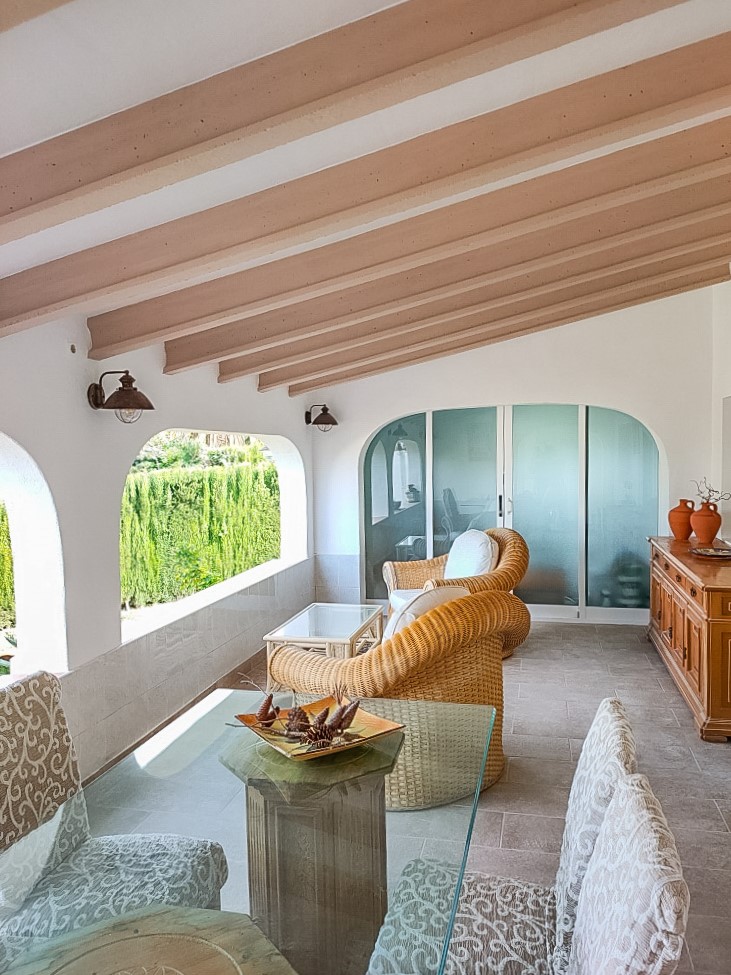 Naya cubierta con vigas de madera a la vista, zona de comedor con mesa de cristal y sillones de mimbre. Los arcos tradicionales ofrecen luz natural y vistas a la vegetación.