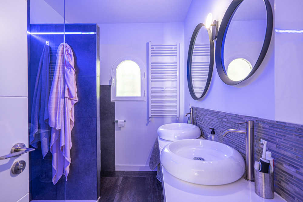 Baño moderno con lavabo doble de seno circular, espejos redondos y ducha italiana con iluminación LED azul y azulejos oscuros.