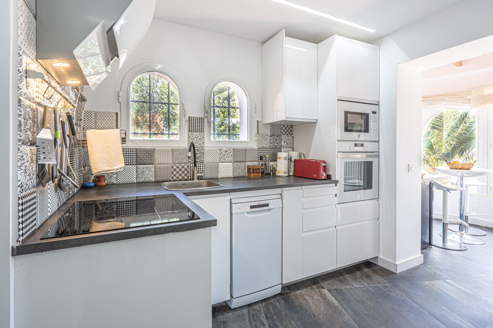 Cocina moderna con armarios blancos brillantes, encimera oscura y salpicadero de azulejos hidráulicos. Incluye electrodomésticos integrados y ventanas de arco con vistas al exterior.