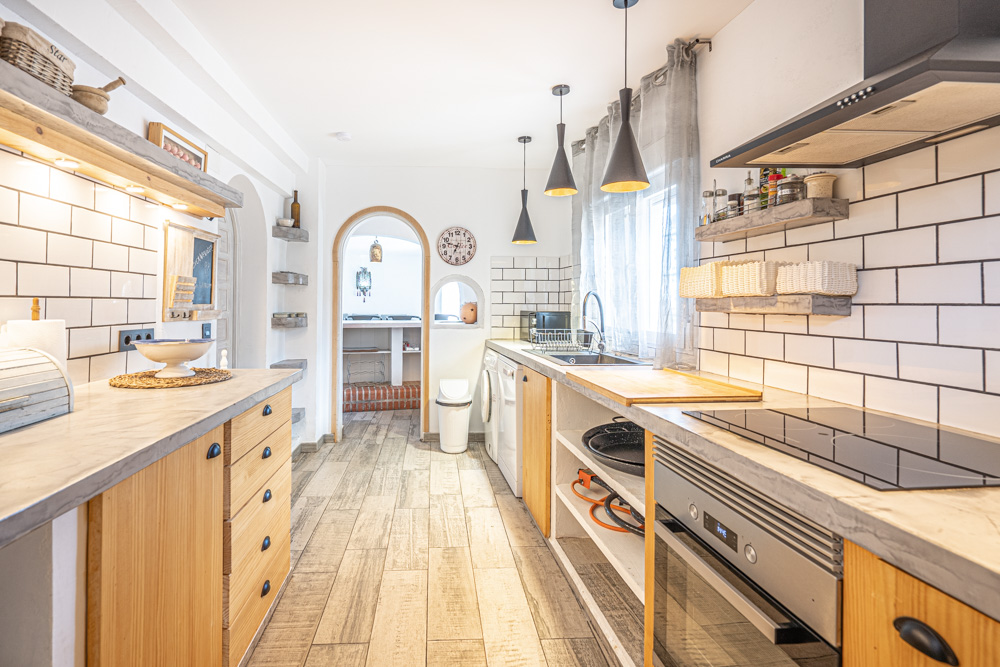 Cocina rústica con encimeras grises, muebles de madera y paredes de azulejo blanco. Incluye electrodomésticos integrados, iluminación colgante y un arco que conecta con el comedor.