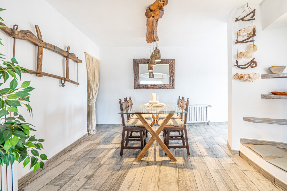 Comedor de diseño rústico con mesa de cristal y base de madera, iluminado por una viga natural tallada y decorado con un gran espejo de marco artesanal sobre suelos cerámicos.