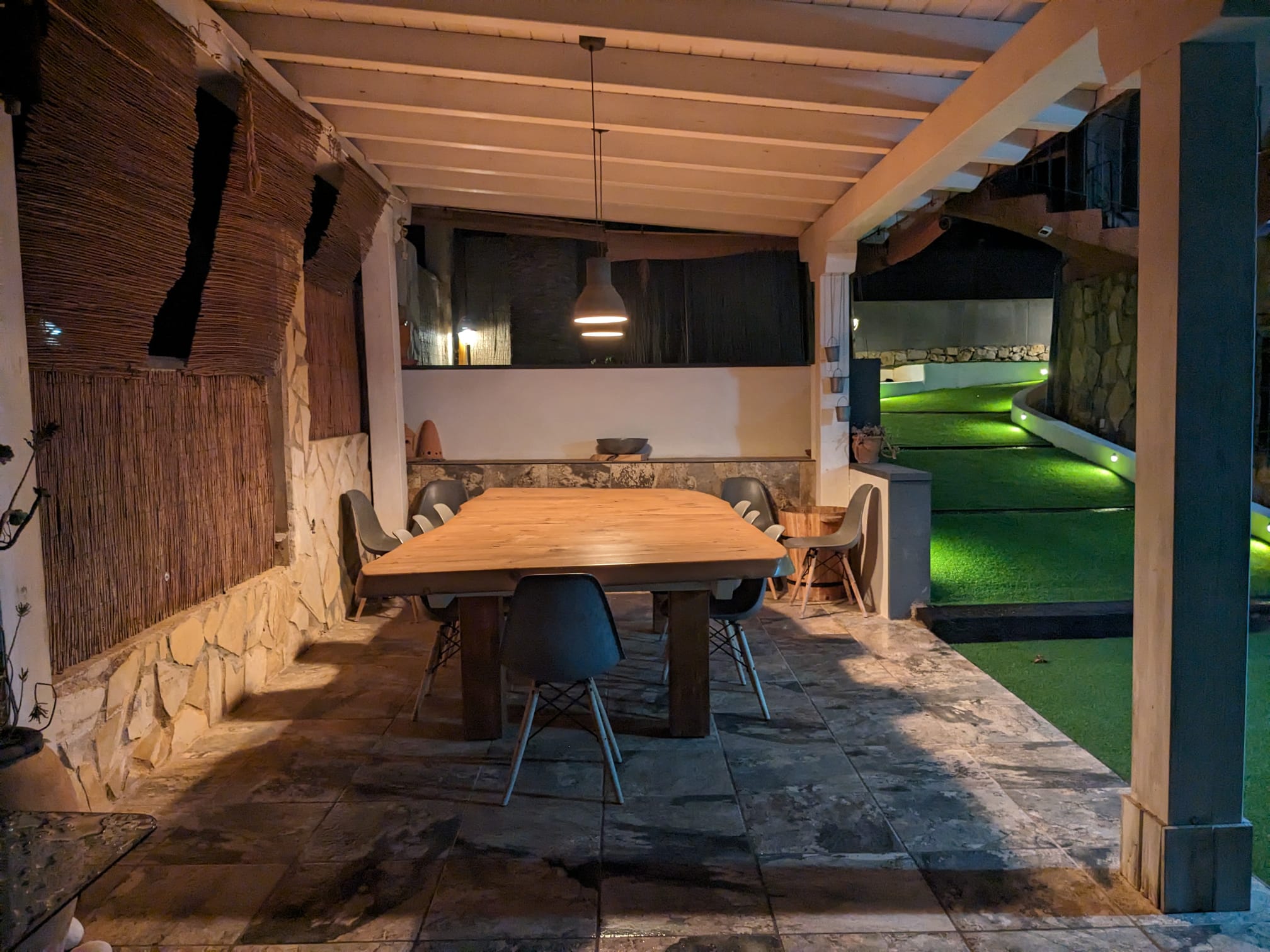 Comedor exterior bajo pérgola con mesa de madera maciza, suelos de piedra natural e iluminación ambiental nocturna junto al jardín.