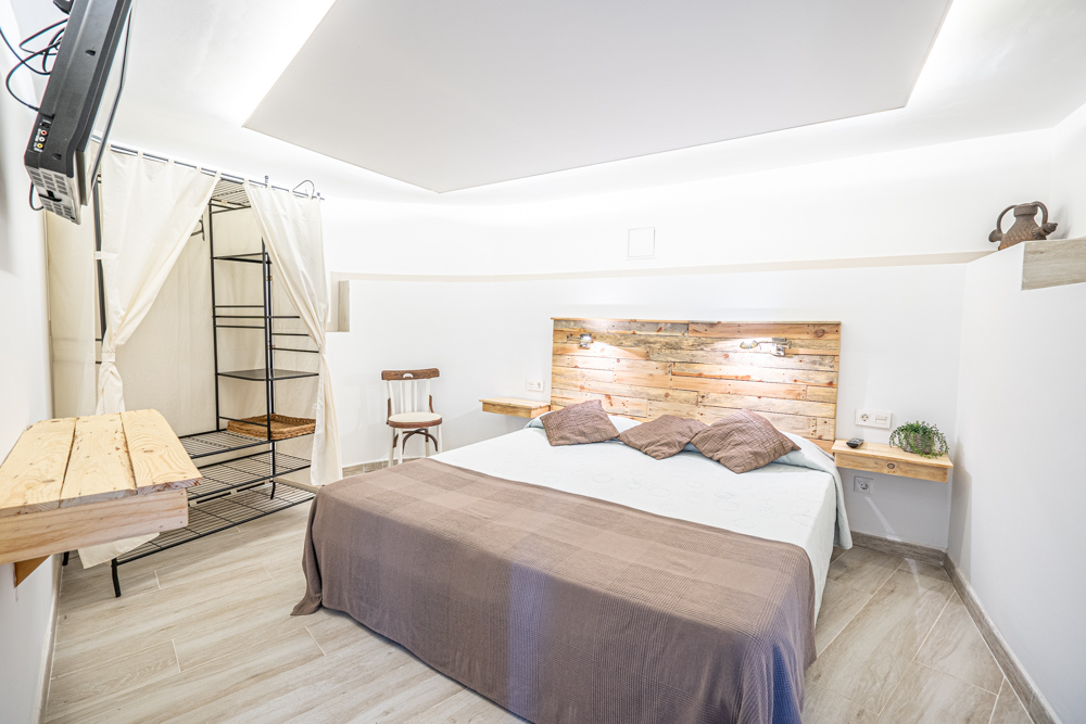 Dormitorio con cabecero de madera rústica, mesitas flotantes a juego, iluminación indirecta en el techo y un práctico armario abierto con cortinas blancas.