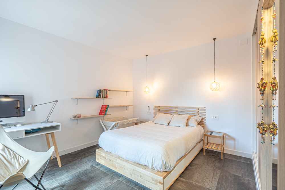 Dormitorio moderno con cama de madera, zona de escritorio integrada y estanterías flotantes. Destacan las lámparas colgantes y el suelo de gres porcelánico oscuro.