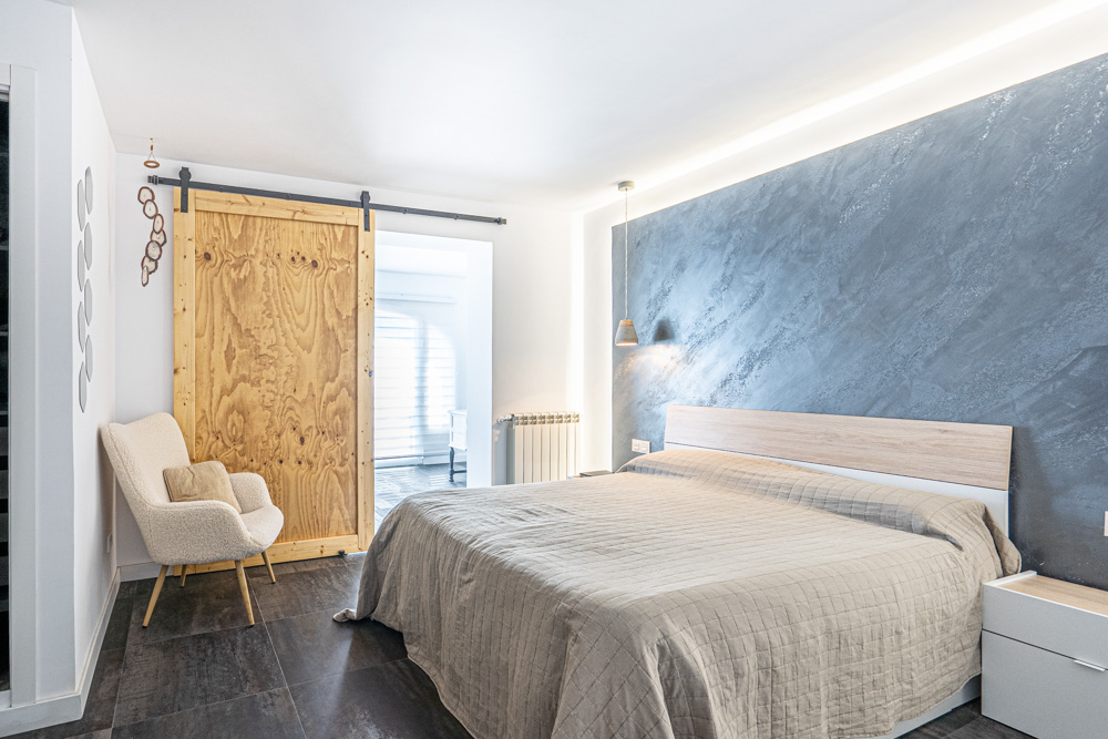 Dormitorio moderno con pared de acento azul texturizada e iluminación LED indirecta, puerta corredera de madera natural y suelos de gres oscuro.
