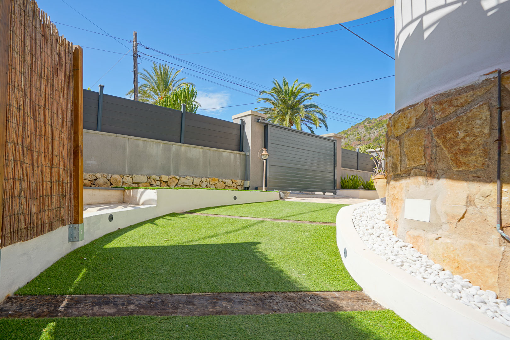 Entrada exterior con césped artificial, traviesas de madera y muros de piedra natural. Cuenta con un portón corredero moderno y bordes de grava blanca decorativa.