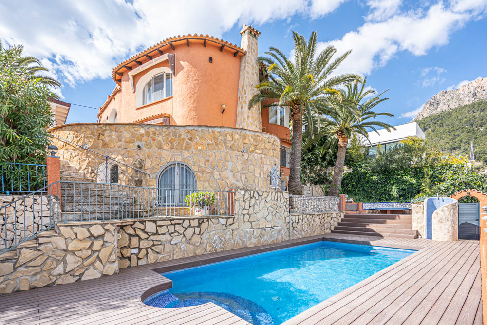 Exterior de la villa con piscina privada, rodeada de tarima y muros de piedra natural, bajo un entorno de palmeras y vistas a la montaña.