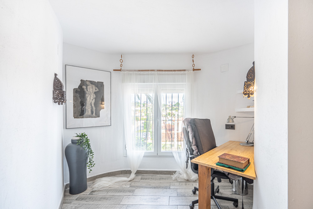Rincón de estudio con escritorio de madera, suelo cerámico de efecto madera y ventana con rejas de seguridad que permite la entrada de luz natural.