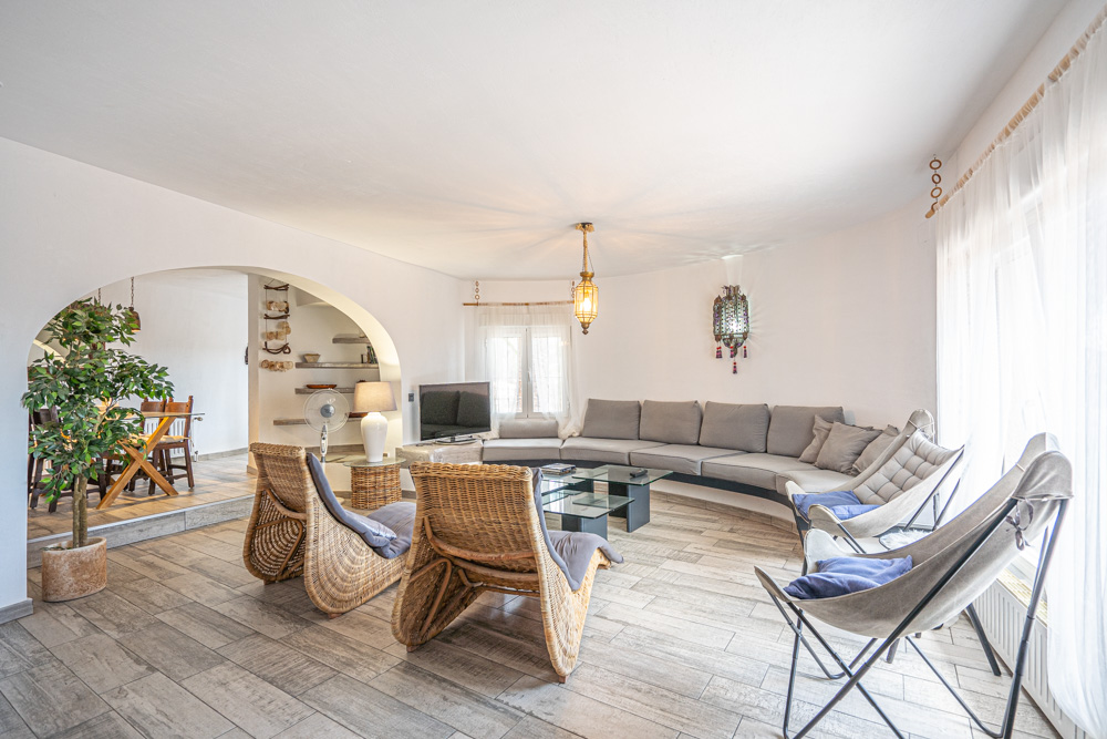 Salón luminoso con sofá curvo integrado, sillones de mimbre y suelos cerámicos de efecto madera. Un arco conecta con el comedor bajo una iluminación de estilo mediterráneo.