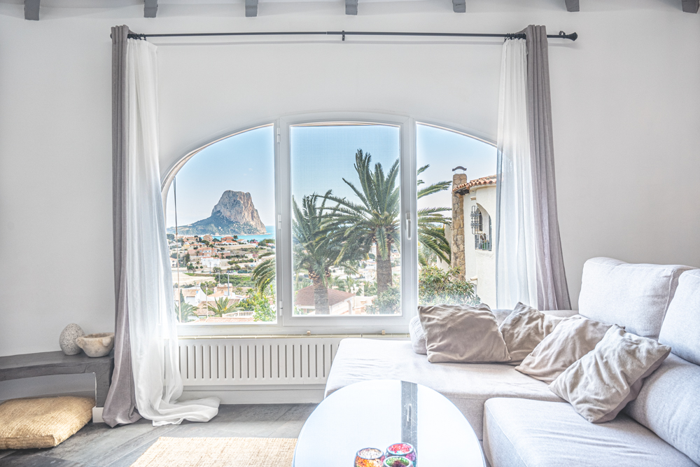 Salón luminoso con sofá rinconera y gran ventanal arqueado que ofrece vistas directas al Peñón de Ifach y las palmeras de Calpe.