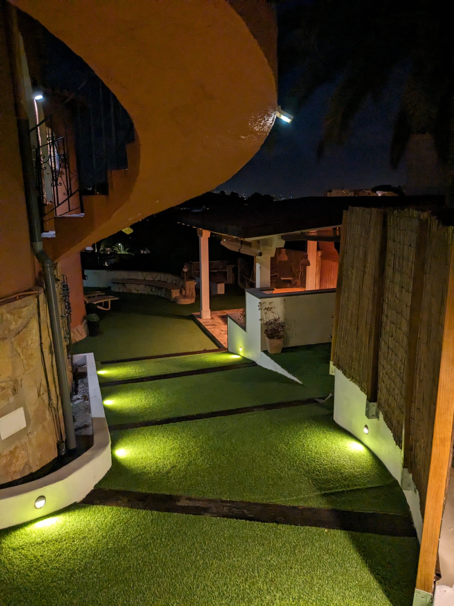 Vista nocturna de los accesos exteriores con iluminación LED integrada, senderos de césped artificial y zona de pérgola techada al fondo.