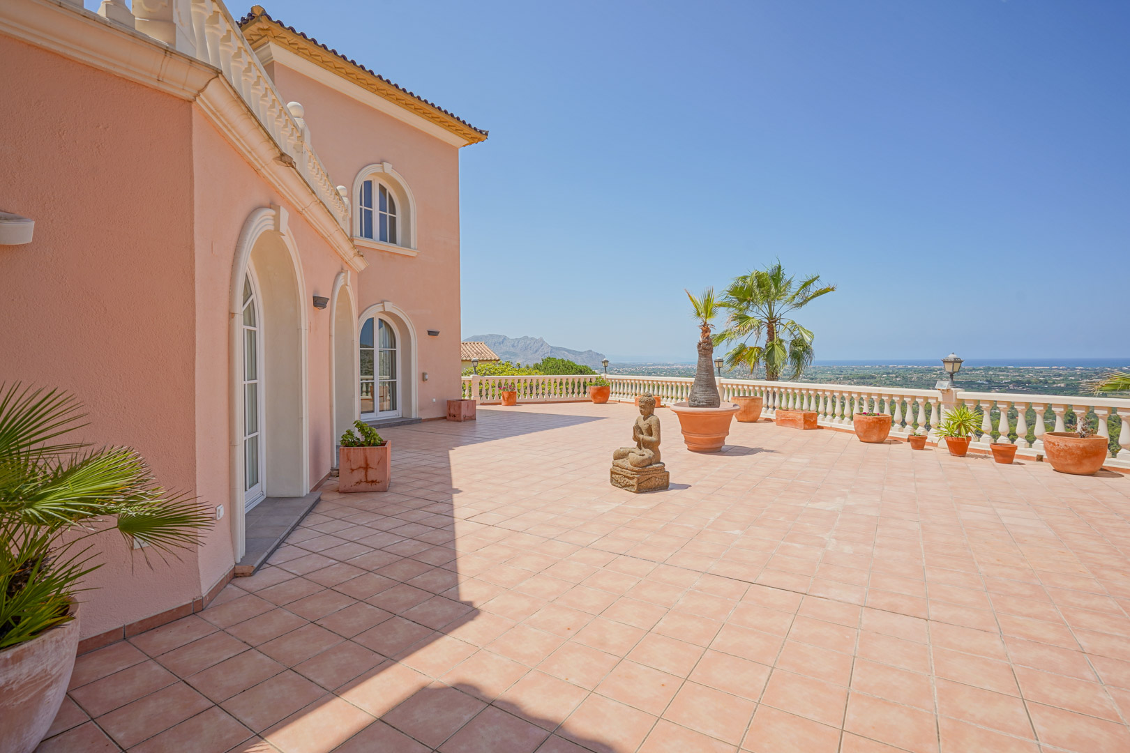 Amplia terraza con pavimentos cerámicos y balaustrada clásica que ofrece vistas panorámicas al mar. La fachada destaca por sus ventanales en arco y decoración mediterránea.