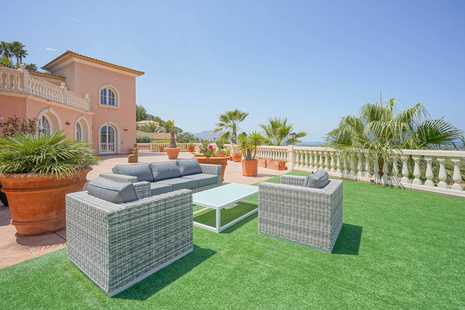Amplia terraza con zona de estar de mimbre gris sobre césped artificial, rodeada de macetas de terracota y vistas despejadas a la costa y montaña.