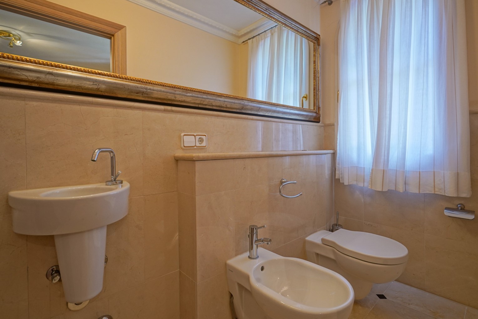 Baño de cortesía revestido en mármol crema, equipado con lavabo suspendido, bidé e inodoro. Incluye un gran espejo con marco decorativo y luz natural filtrada por cortinas.