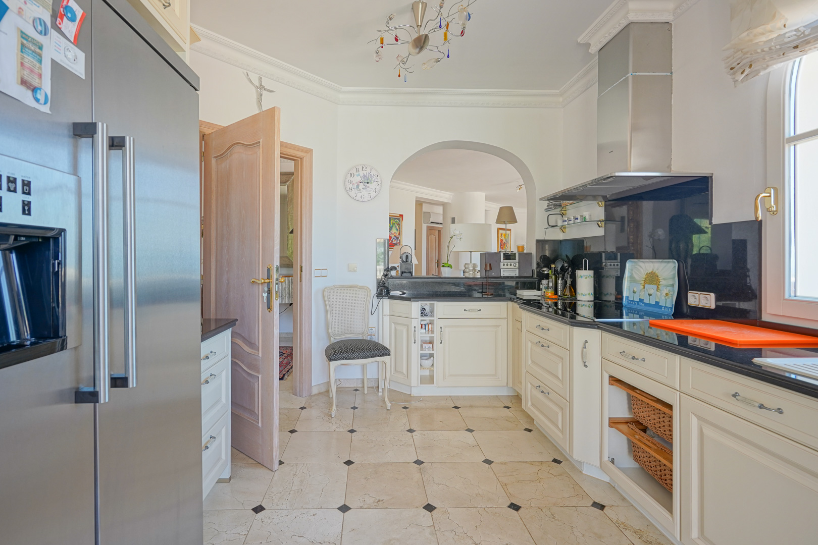 Cocina equipada con mobiliario en tono crema, encimeras de granito negro y suelo de mármol. Incluye electrodomésticos de acero inoxidable y un arco que conecta con la zona de estar.