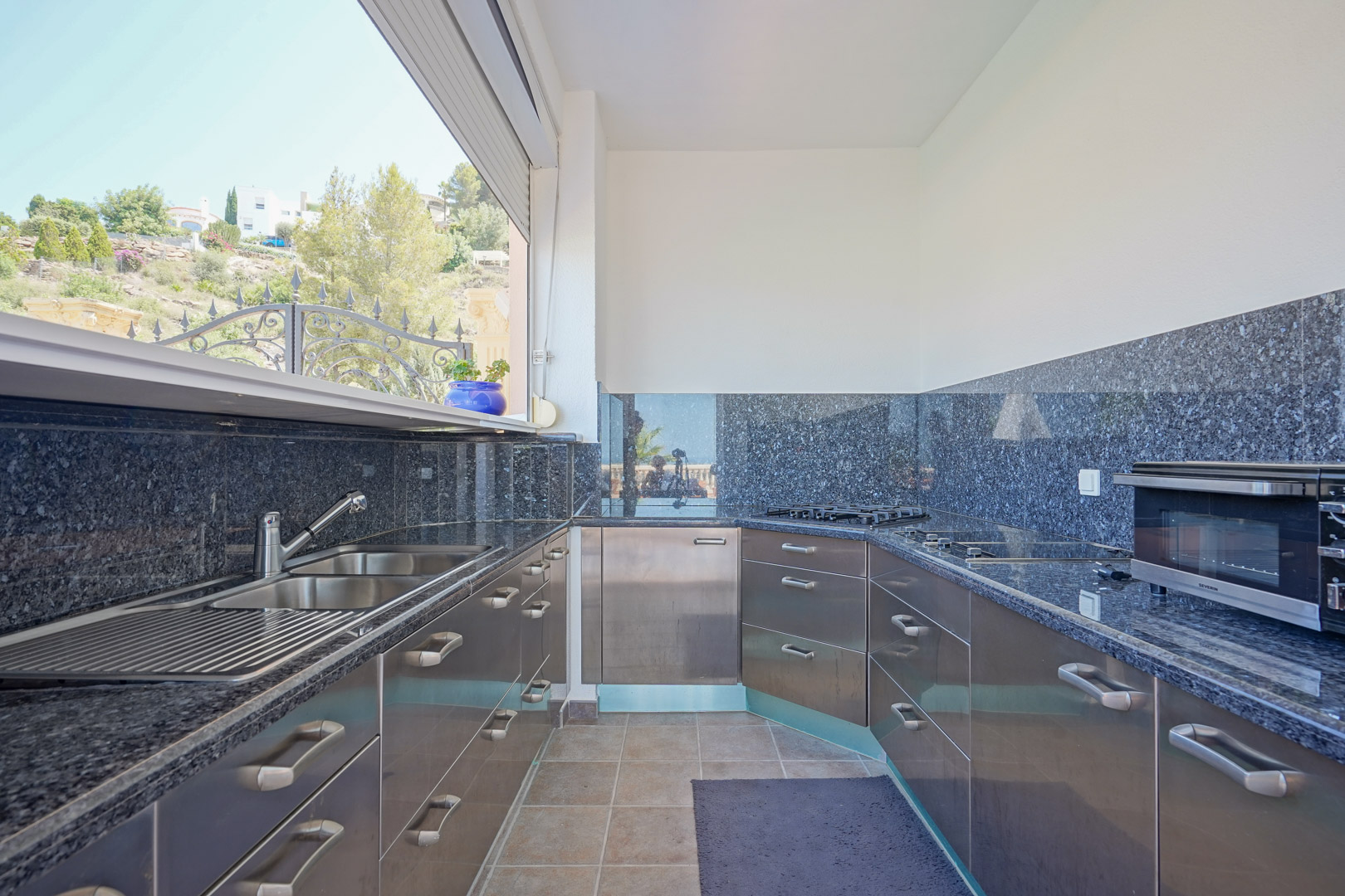 Cocina exterior con encimeras de granito oscuro, armarios de acero inoxidable y fregadero doble, situada frente a un ventanal con vistas al entorno natural de La Sella.