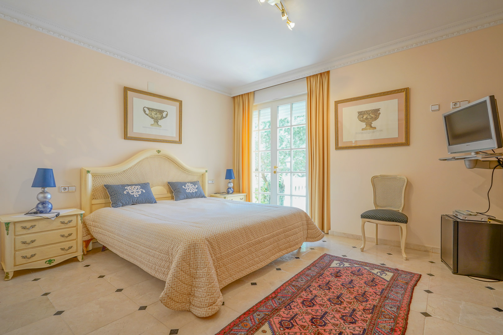 Dormitorio luminoso con suelos de mármol y detalles clásicos. Incluye acceso directo al exterior mediante puertas acristaladas y mobiliario de estilo tradicional en tonos crema.