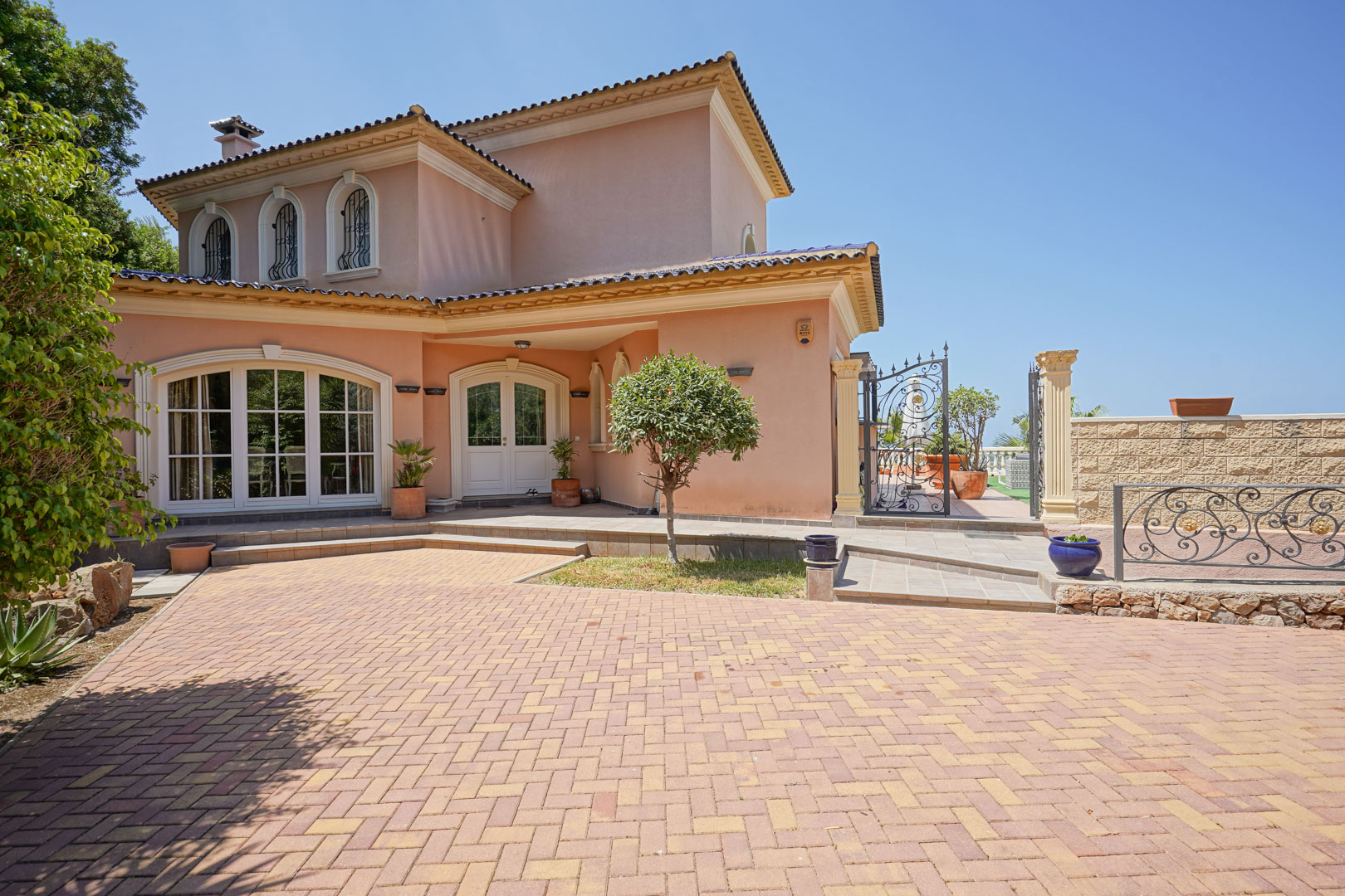Fachada de la villa con entrada principal, ventanales arqueados y amplio acceso pavimentado. Incluye detalles de forja en puertas y ventanas bajo un cielo despejado.