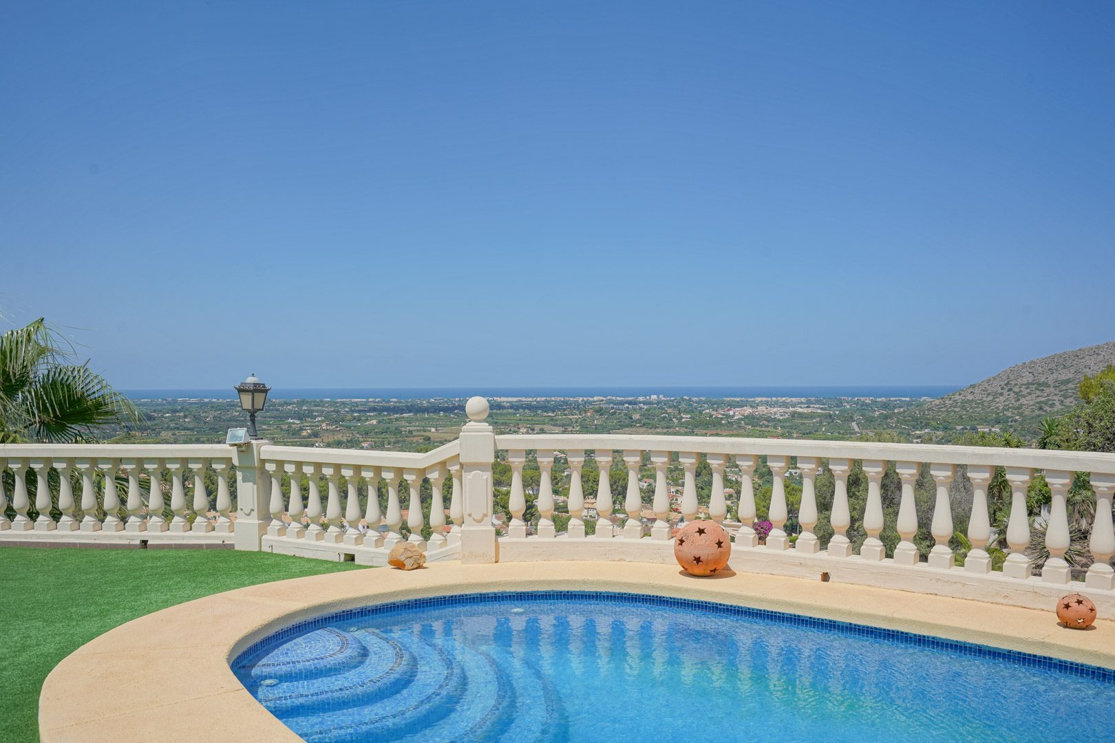 Piscina curva con gresite azul y balaustrada clásica, ofreciendo vistas panorámicas despejadas hacia el valle y el mar Mediterráneo bajo un cielo despejado.