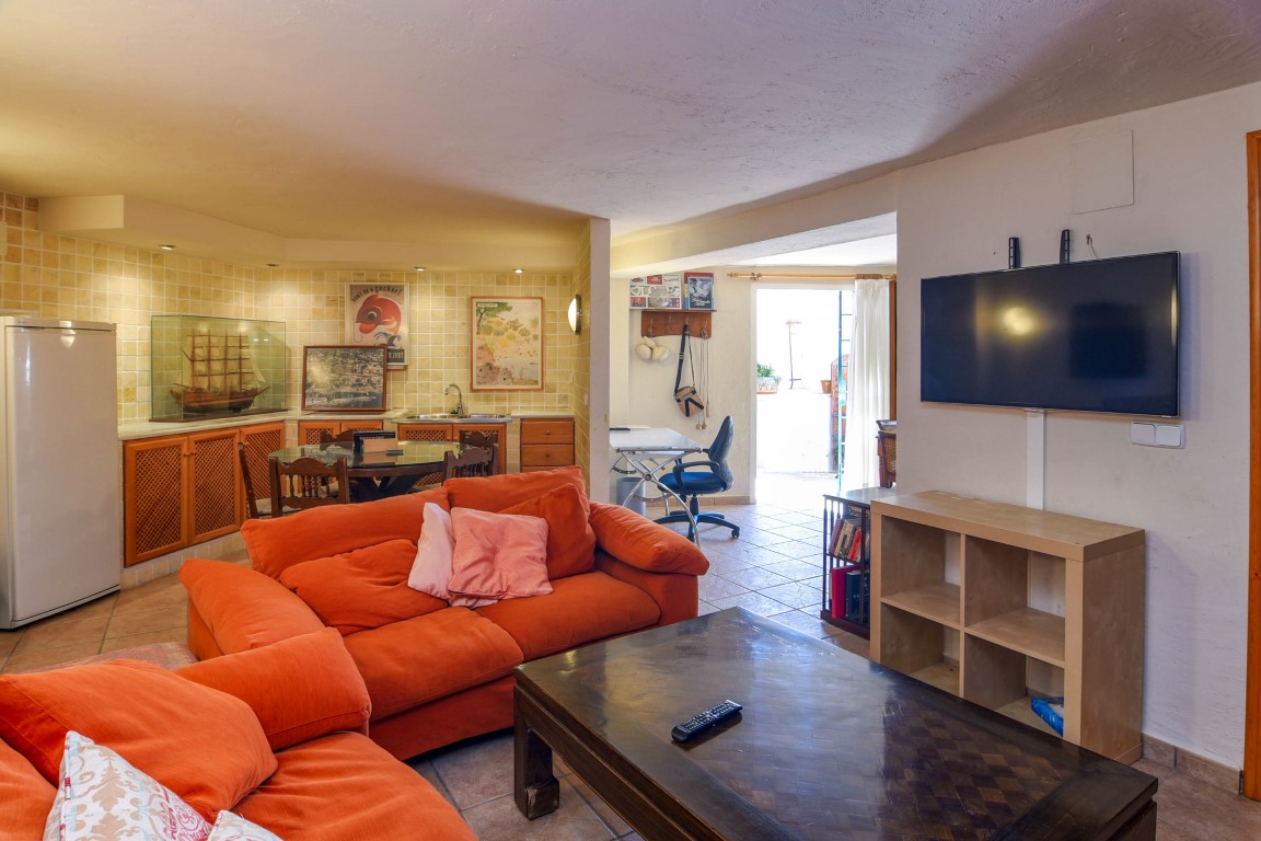 Sala de estar polivalente con amplios sofás naranjas, zona de oficina y una cocina integrada con acabados en madera y azulejos rústicos.