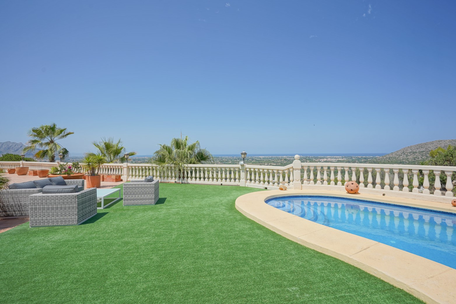 Terraza exterior con piscina de borde curvo, zona de césped artificial y mobiliario de ratán sintético con vistas panorámicas al mar y al valle de La Sella.