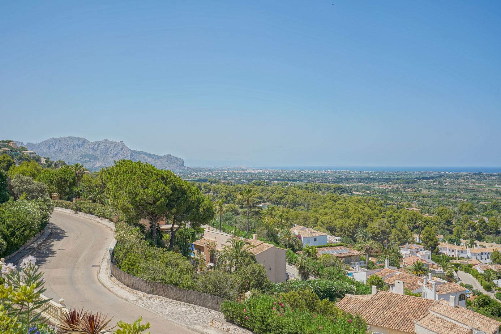Vistas panorámicas desde la zona elevada de La Sella, mostrando la costa mediterránea, el valle arbolado y el macizo del Montgó bajo un cielo despejado.