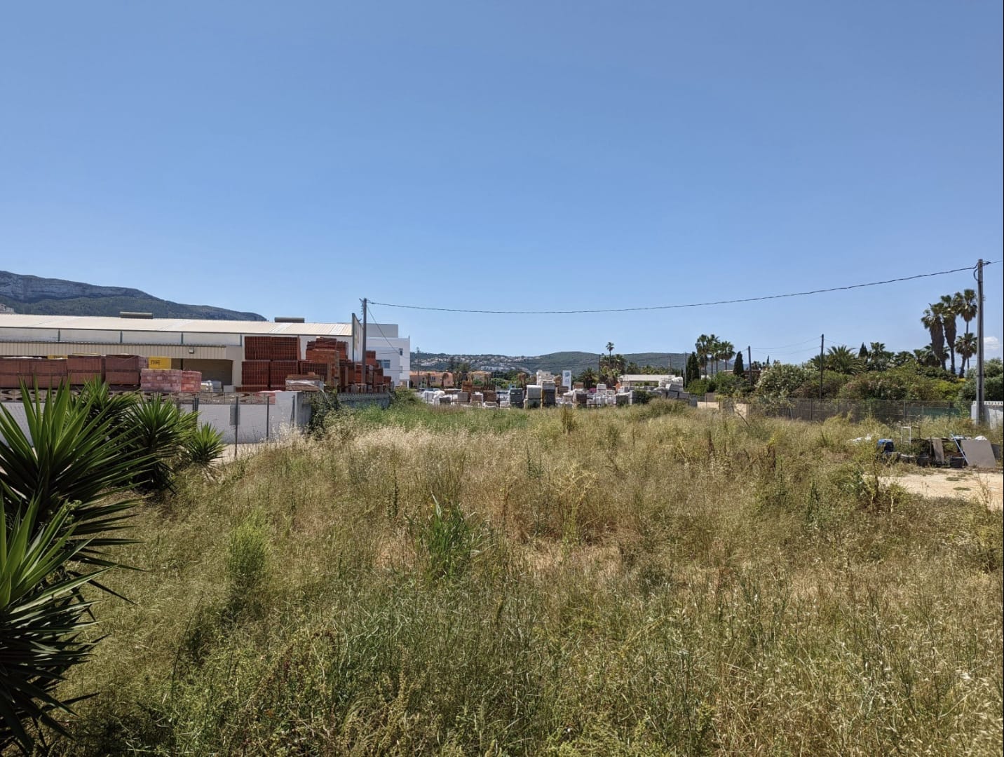 Parcela llana con vegetación autóctona y vistas al Montgó. El terreno se encuentra delimitado y situado junto a una zona de servicios bajo un cielo despejado.