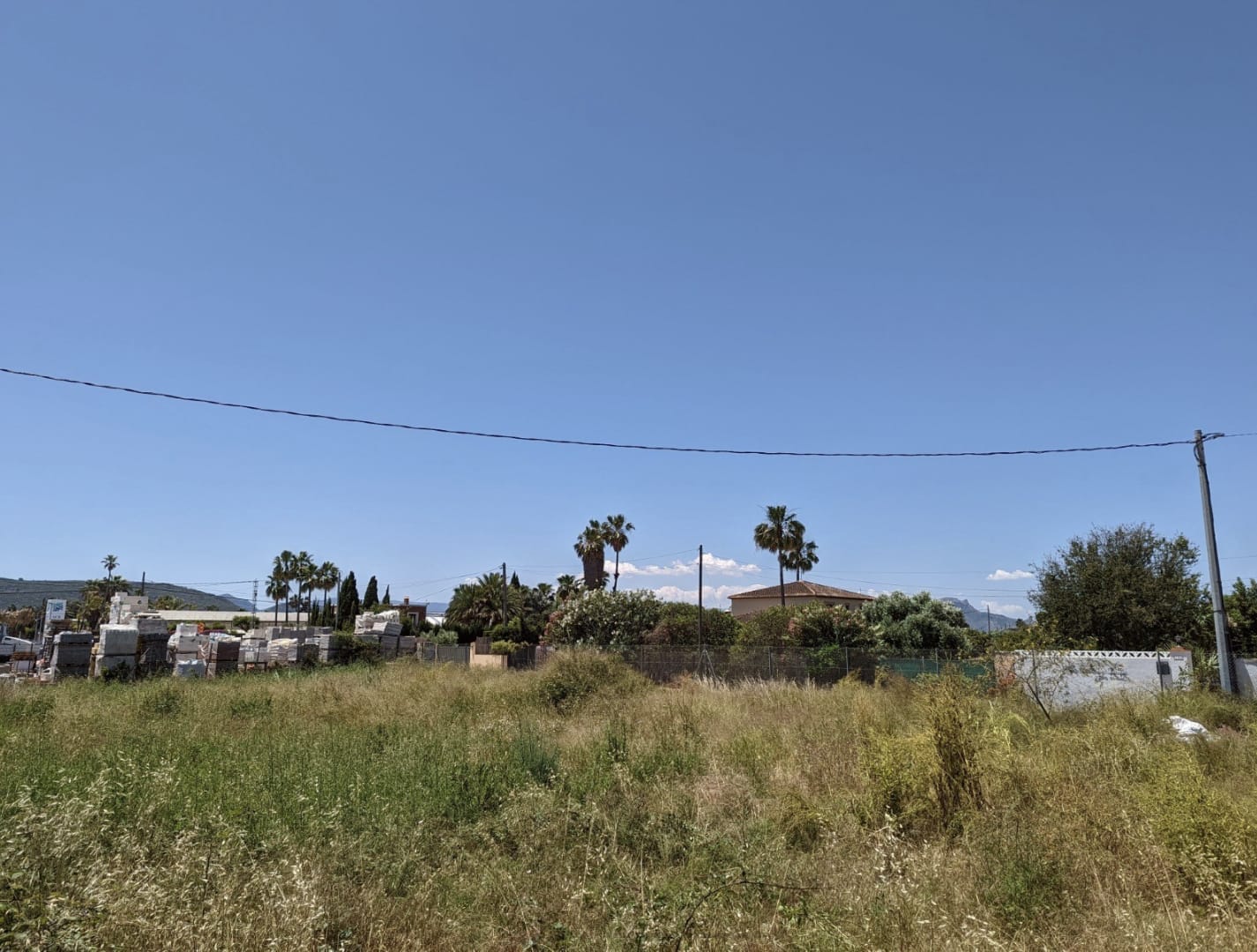 Terreno llano con vegetación baja y materiales de construcción acopiados. Vista despejada hacia palmeras y edificaciones residenciales bajo un cielo despejado.
