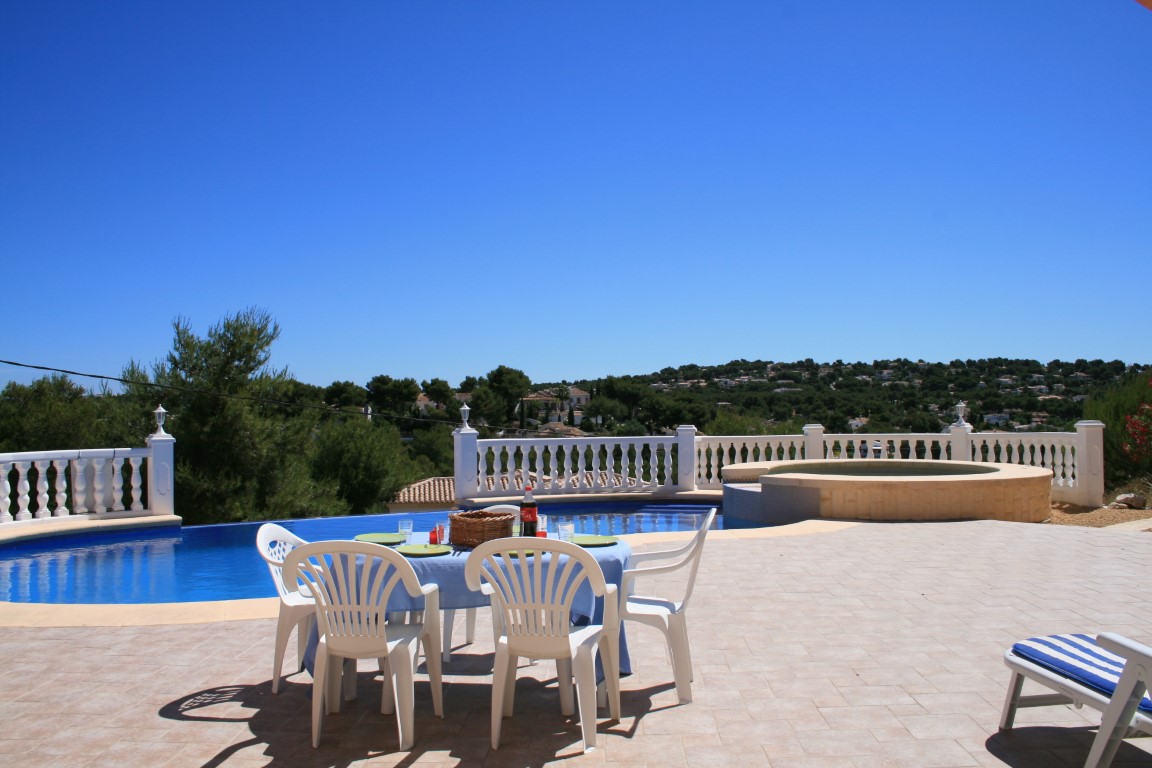 Amplia terraza exterior con piscina de forma curva, jacuzzi circular integrado y zona de comedor con vistas panorámicas a las colinas de Jávea.
