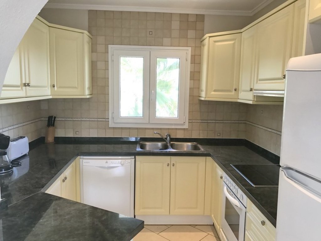 Cocina totalmente equipada con armarios en tono crema, encimeras de granito oscuro y fregadero doble de acero inoxidable bajo una ventana con luz natural.