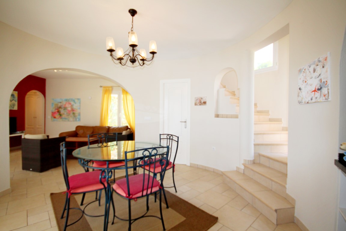 Comedor de planta abierta con mesa de cristal y sillas de hierro forjado, conectado al salón mediante un arco y situado junto a la escalera de gres de tonos claros.