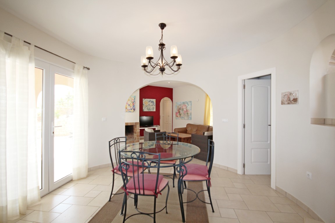 Comedor luminoso con mesa de cristal y sillas de forja, conectado mediante un arco al salón que cuenta con chimenea y una pared de acento roja.