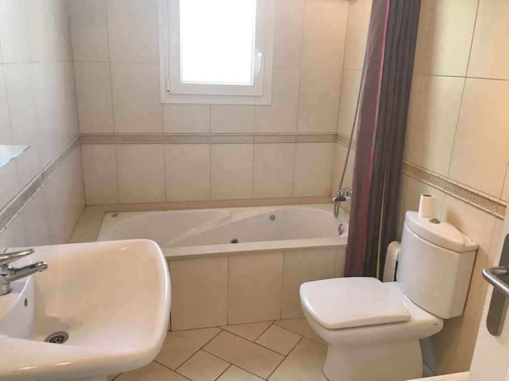 Cuarto de baño con bañera de hidromasaje integrada, lavabo de pedestal blanco e inodoro. Presenta paredes alicatadas en tonos crema con cenefa decorativa y luz natural.