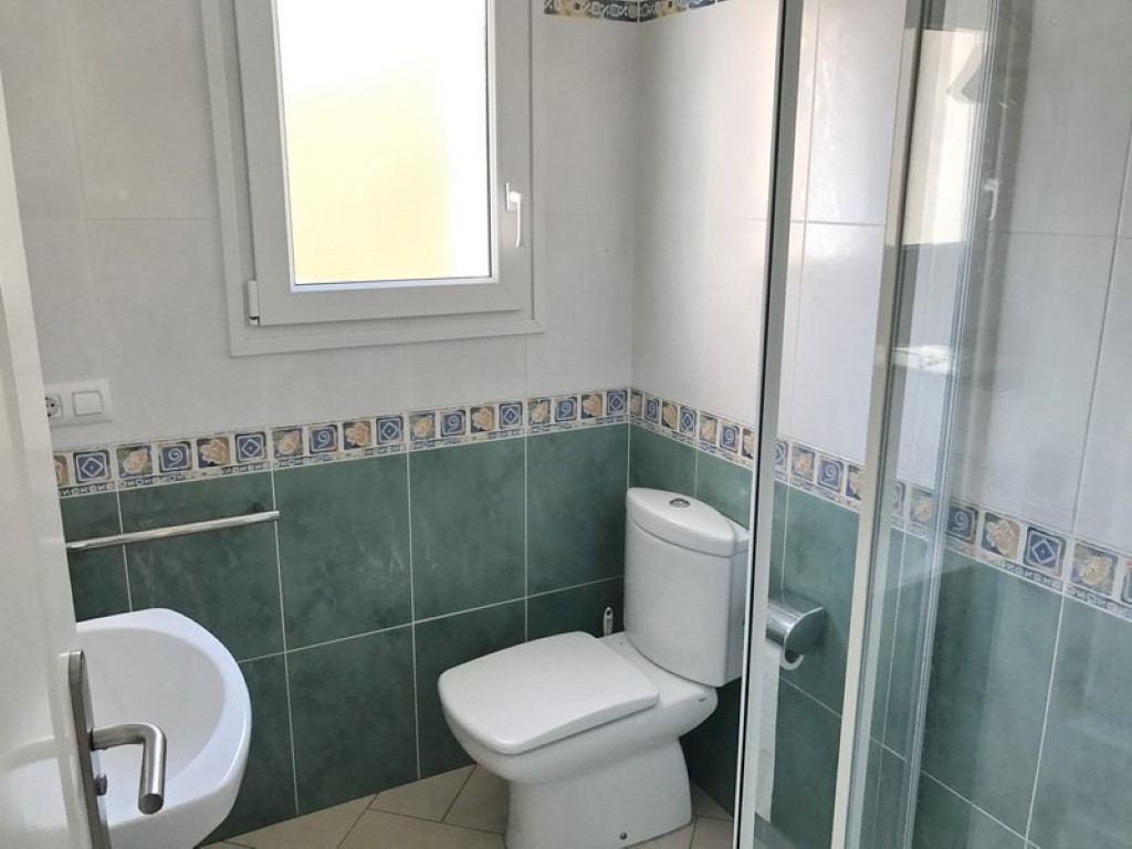 Cuarto de baño con sanitarios blancos, azulejos en tonos verdes y blancos con cenefa decorativa, y una ducha con mampara de cristal junto a la ventana.