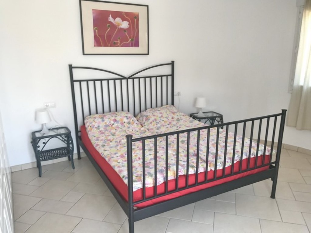 Dormitorio con cama de forja negra, mesitas de noche de mimbre y cristal, y suelos de baldosa clara. Dispone de luz natural y una decoración de pared minimalista.