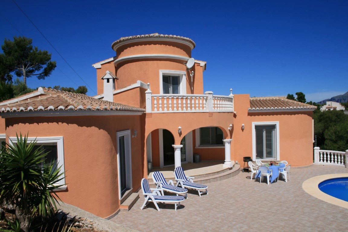 Exterior de villa mediterránea con fachada terracota, torreón circular, amplia terraza de gres con tumbonas y zona de piscina bajo un cielo despejado.