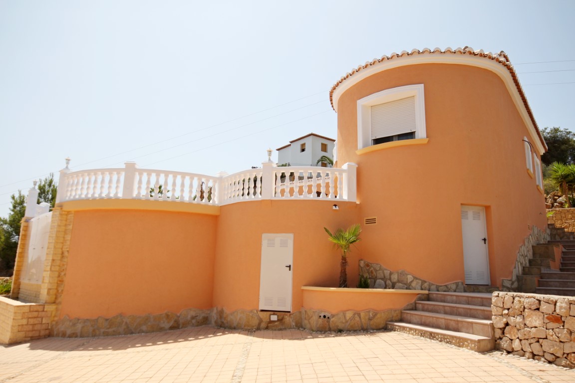 Fachada exterior curva en tono terracota con balaustrada blanca clásica, base de piedra natural y patio pavimentado bajo el sol mediterráneo.