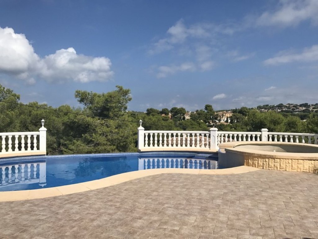 Piscina curva con jacuzzi integrado rodeada de una terraza de gres y balaustrada blanca, ofreciendo vistas despejadas a la vegetación mediterránea de Jávea.
