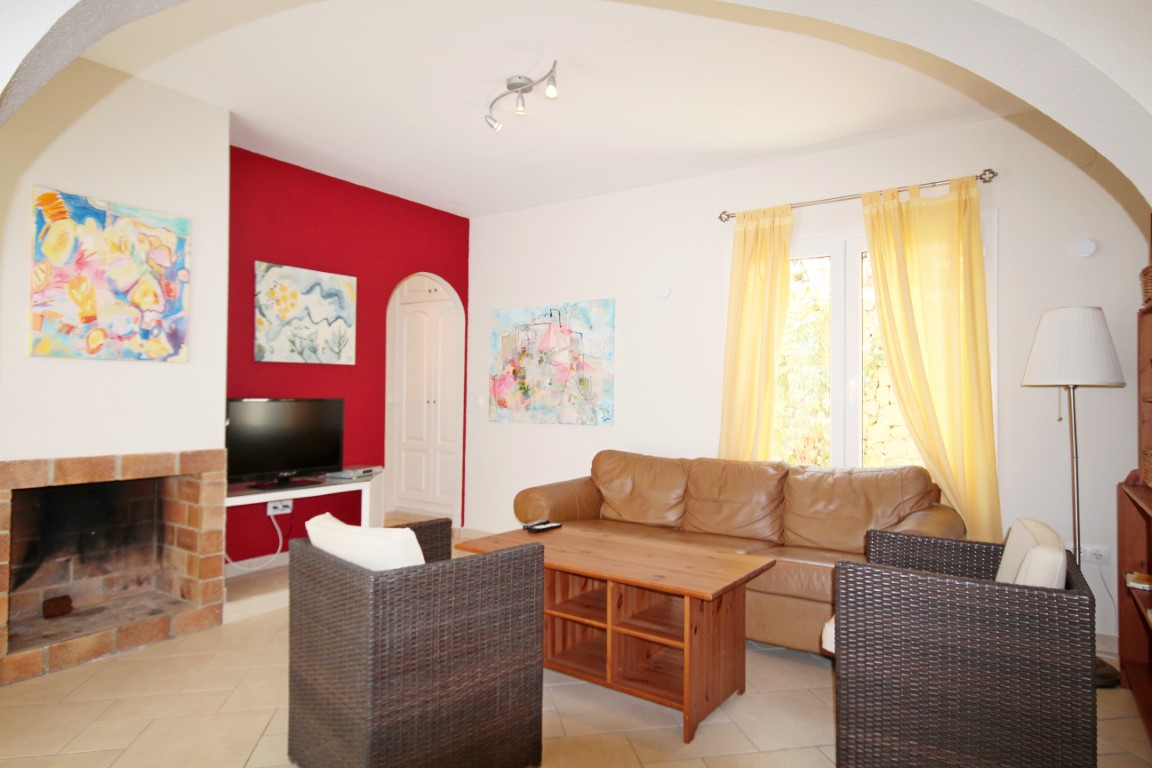 Salón con chimenea de ladrillo, pared de acento roja y sofá de cuero. Dispone de sillones de mimbre, mesa de centro de madera y luz natural a través de cortinas amarillas.