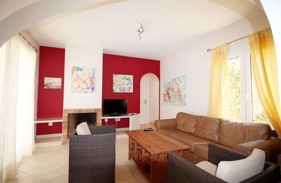 Salón con pared de acento roja, chimenea de ladrillo tradicional y mobiliario de cuero marrón junto a sillones de mimbre oscuro y suelos cerámicos claros.