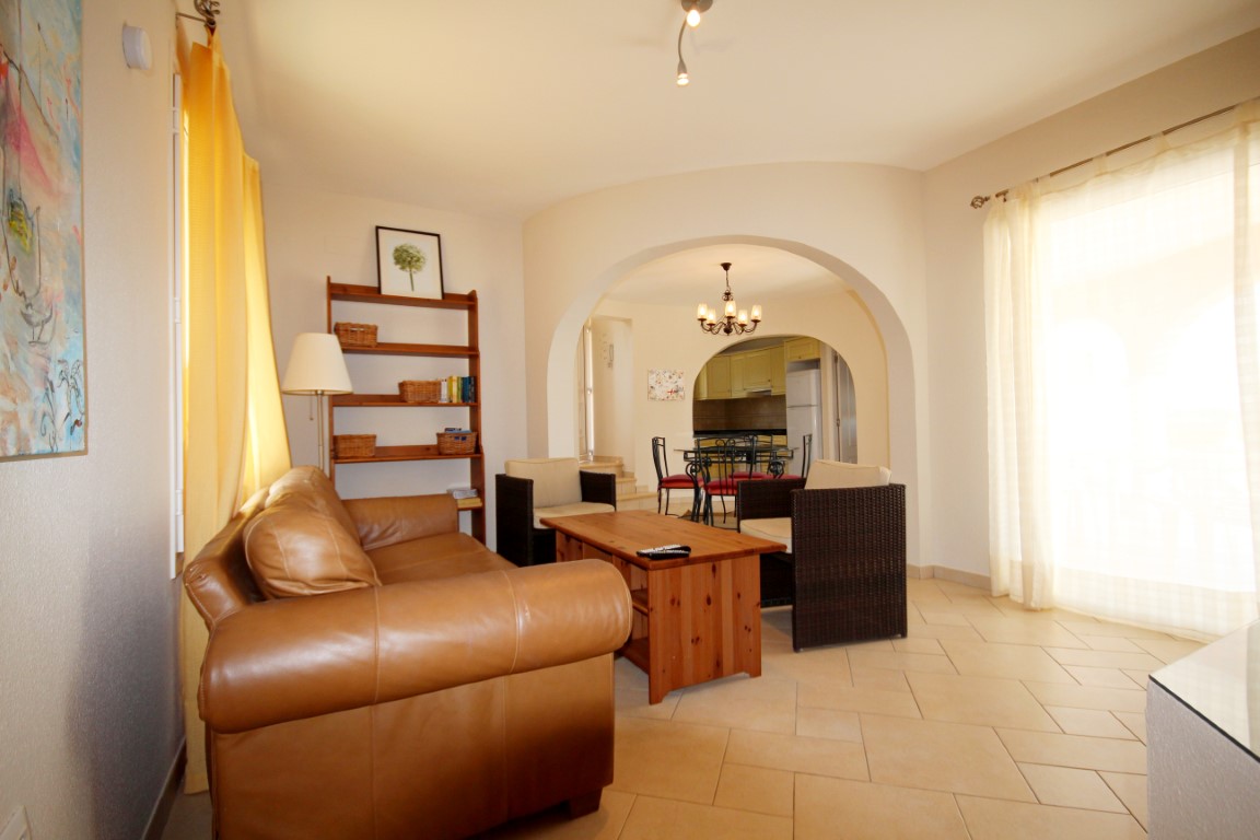 Salón con sofá de cuero marrón, mesa de centro de madera y sillones de mimbre. Un arco arquitectónico comunica la zona de estar con el comedor y la cocina al fondo.