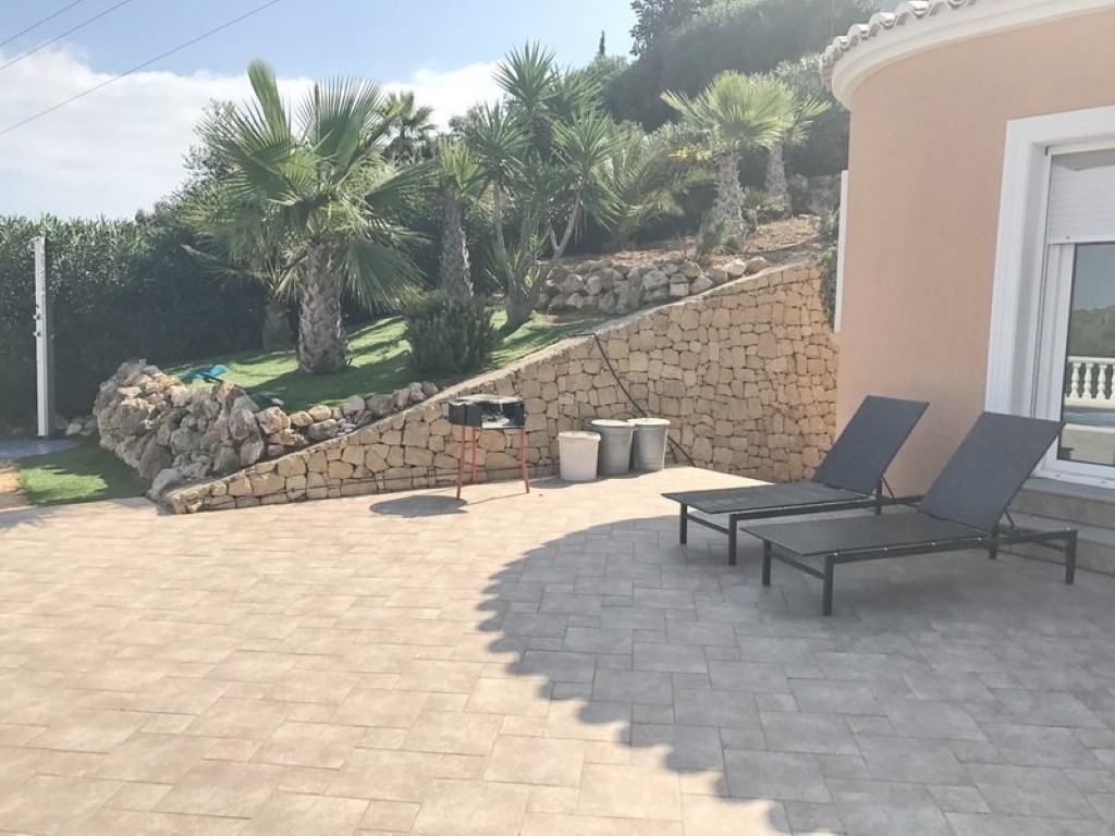 Terraza exterior pavimentada con tumbonas negras, muro de piedra natural y jardín mediterráneo con palmeras junto a la fachada de la villa.
