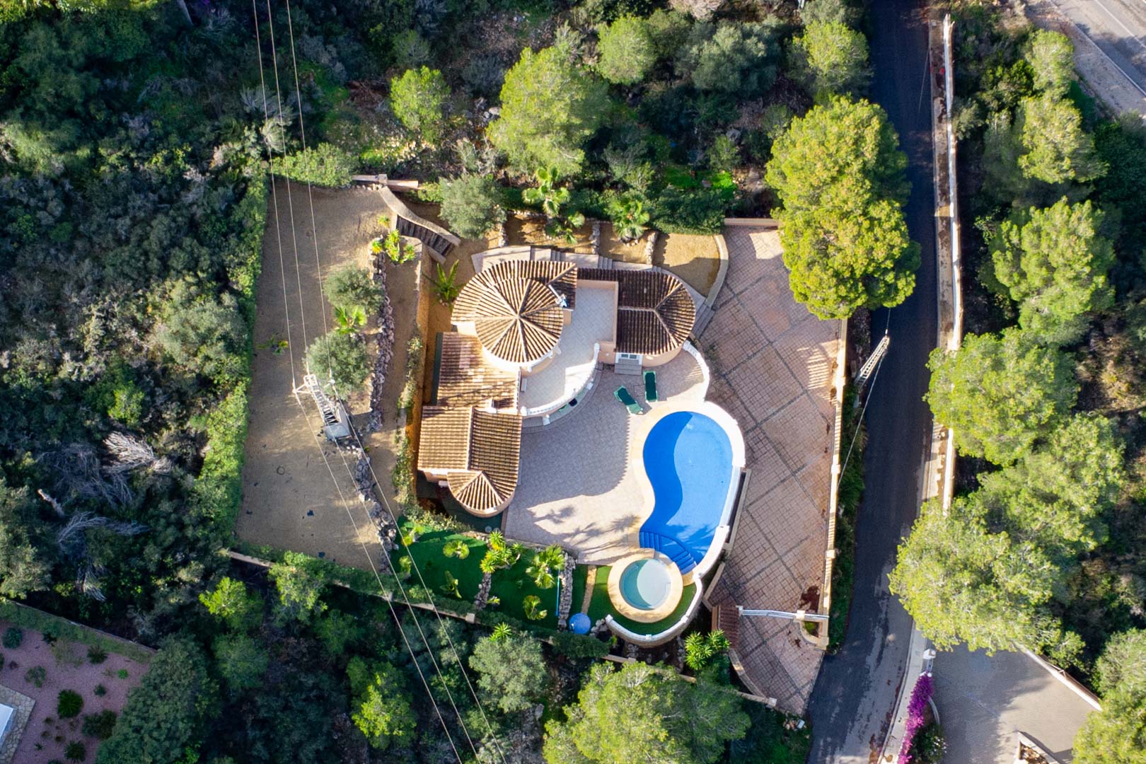 Vista aérea de la villa que muestra los tejados de teja cerámica, la piscina en forma de riñón y el jacuzzi circular, rodeados de un denso bosque mediterráneo.